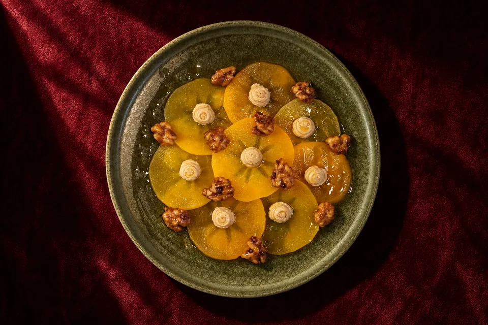 El mismo carpaccio de caqui sobre terciopelo granate con sombras marcadas. Foto de estudio creada con nuestro motor de inteligencia artificial.