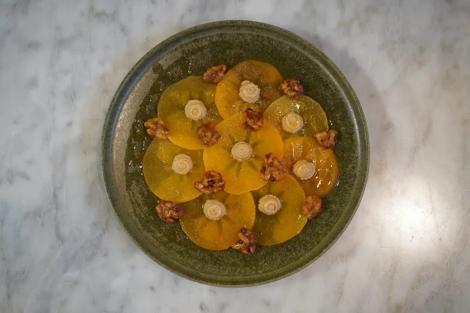 Carpaccio de caqui con nueces garrapiñadas y rosetas de crema en un plato verde, desde arriba sobre mármol: la foto de partida.